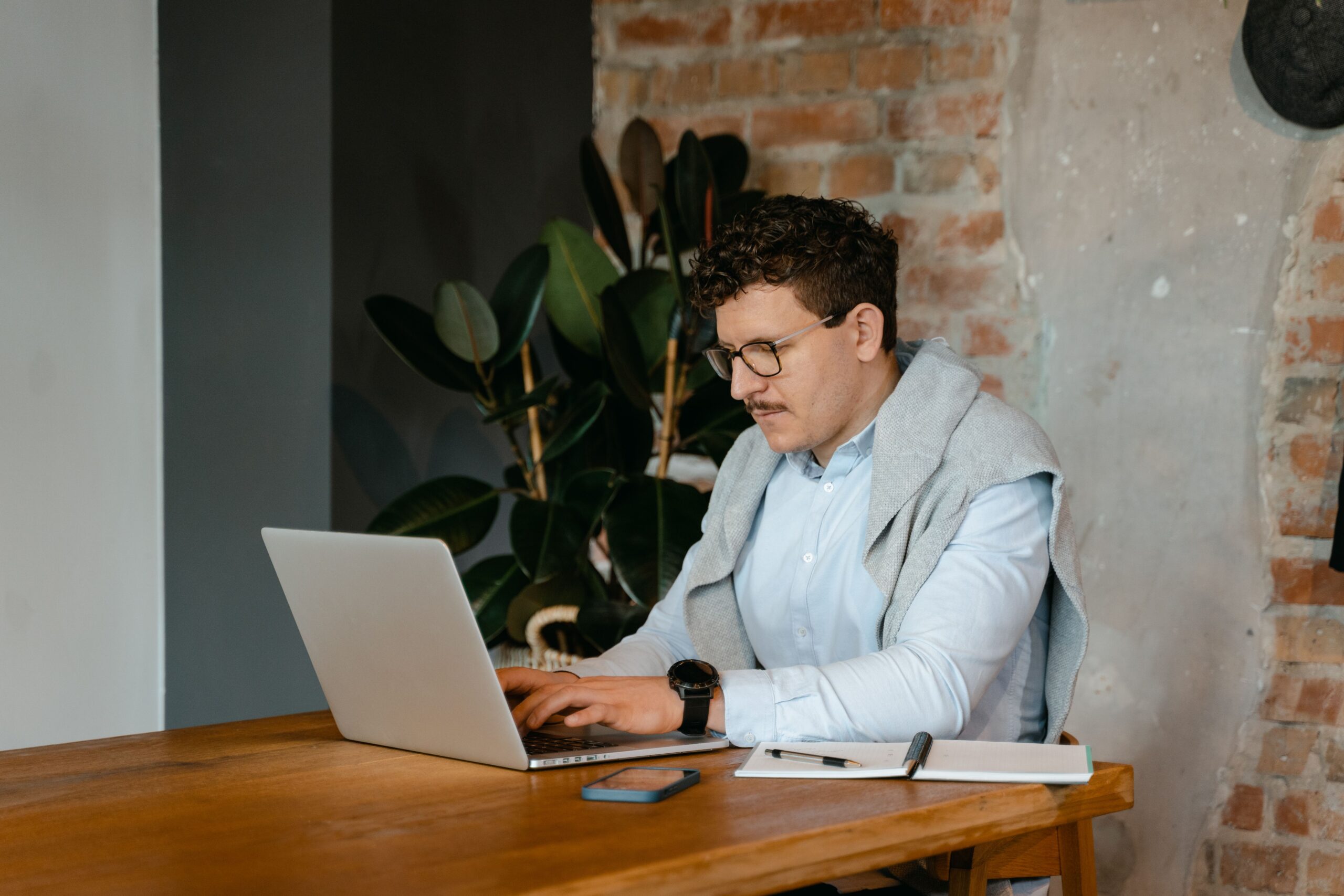 A person working at a laptop beside a notebook and plant.